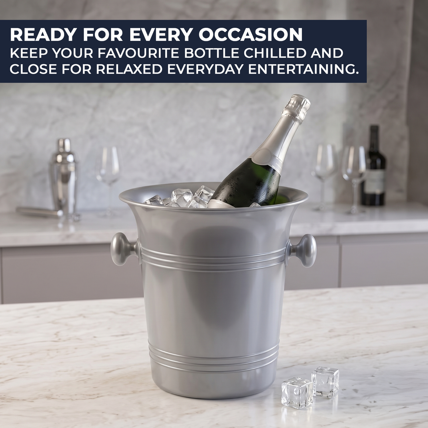 A silver metal champagne bucket filled with ice and a bottle of champagne, set on a marble countertop in a kitchen-like setting. A dark blue text banner at the top reads, 'READY FOR EVERY OCCASION KEEP YOUR FAVOURITE BOTTLE CHILLED AND CLOSE FOR RELAXED EVERYDAY ENTERTAINING.