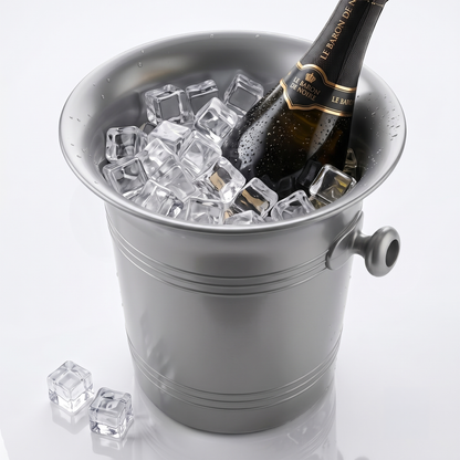 A silver metal champagne bucket filled with clear ice cubes, with a dark champagne bottle resting inside. Two clear ice cubes sit on the reflective surface beside the bucket.